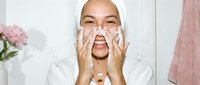 Young Asian woman applying facial cleanser with foam on her hands in modern bathroom during morning skincare routine for healthy glowing skin care regimen.