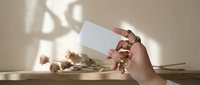 Female hand holding blank white card with dried flowers and soft natural lighting creating elegant shadows on beige background for branding mockup.