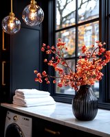 Elegant laundry room interior with coral flowers in black vase, stacked white towels, pendant lights, and window view. Modern minimalist design with autumn accents.