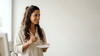 Young woman with long dark hair and South Asian ethnicity holding a plate, wearing casual beige clothing in bright interior space for lifestyle and wellness content.