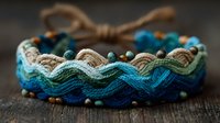 Handcrafted woven bracelet with wavy pattern in shades of blue and beige, adorned with small turquoise and copper beads on rustic wooden surface.