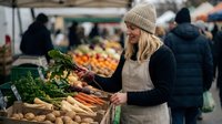 Young blonde woman in beige knit hat and apron selecting fresh vegetables at outdoor farmers market stall with organic produce display for healthy lifestyle.