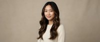 Young Asian woman with long wavy brown hair wearing white sweater smiling at camera against neutral beige background for portrait photography.