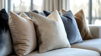 Cozy arrangement of decorative throw pillows in neutral tones of beige, gray and brown on modern sofa near window, creating relaxing home atmosphere.