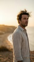 Handsome middle-aged Caucasian man with curly hair in linen shirt standing on coastal cliff during golden hour sunset with ocean waves in background.