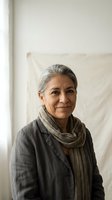 Confident mature Hispanic woman with gray hair wearing linen jacket and knit scarf smiling warmly in bright modern interior space for lifestyle content.