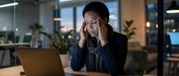 Stressed Asian businesswoman experiencing headache while working late at laptop in modern office environment during evening hours.