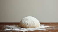 Fresh raw dough ball dusted with flour on wooden surface against neutral background, ready for baking bread or pizza preparation.