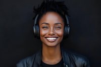 Young African American woman with dreadlocks wearing headphones and leather jacket smiling joyfully against dark background for music and lifestyle content.