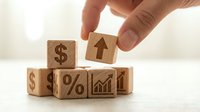Hand placing wooden cube with upward arrow onto stack of blocks featuring dollar signs, percentage and chart symbols representing financial growth and investment success.