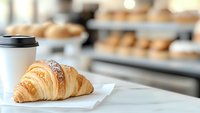 Fresh baked croissant on white paper with takeaway coffee cup in cafe setting. Blurred pastry display case background creates depth and ambiance.