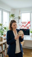 Woman real estate agent holding miniature house model in modern office with rising interest rates chart graphic overlay, representing mortgage rates and property investment concerns.