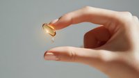 Female hand holding glowing golden vitamin E capsule with oil drop falling against gray background, wellness and skincare concept.