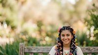 Young woman with long braided hair and colorful beads sitting on wooden bench in sunny garden with blurred green foliage background. Outdoor portrait for lifestyle and fashion content.