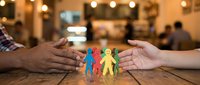 Two hands protecting colorful paper cut family figures on wooden surface with blurred interior background, representing care, unity, insurance, and togetherness for design use.