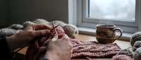 Hands knitting burgundy wool sweater by window with ceramic mug, cozy crafting scene for relaxation and handmade hobby projects at home.