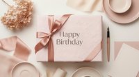 Elegant pink gift box with Happy Birthday inscription, satin ribbon bow, pen, dried flowers, and pastel tableware on white background.
