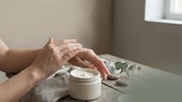 Woman applying moisturizing cream from jar on hands at bathroom vanity. Skincare routine, hand care, beauty treatment, cosmetics application concept.