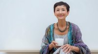 Asian woman with short hair wearing colorful shawl holds white coffee cup while relaxing against white wall, expressing joy and contentment in casual home setting.