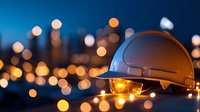 White construction hard hat with glowing light bulb and bokeh city lights background at dusk. Safety equipment for engineering and building projects.