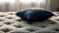 Dark blue pillow resting on beige quilted mattress with city skyline visible through window behind, conveying comfort and relaxation for bedroom design and home decor projects.