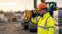 Construction worker in safety gear adjusting hard hat at industrial site during golden hour with heavy machinery in background for workplace safety concepts.
