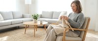 Middle-aged woman with light brown hair, Caucasian ethnicity, sitting in wooden armchair knitting with yarn in bright living room with sofa and natural light for home crafts and leisure.