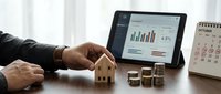 Businessman analyzing financial data on tablet with stacked coins and wooden house model on desk for investment planning and real estate business strategy.