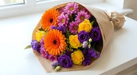 Colorful mixed flower bouquet with orange gerberas, yellow roses and purple blooms wrapped in kraft paper on white surface for gifting occasions.