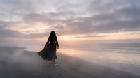 Silhouette of woman in flowing dress walking on misty beach at sunset with dramatic cloudy sky and ocean waves reflecting golden light creating peaceful atmosphere.