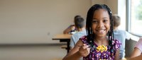 Smiling African American girl eating lunch at school cafeteria, happy child with braids enjoying meal, classmates in background.