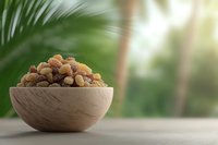 Golden raisins and dark sultanas piled in wooden bowl against tropical palm leaf background, natural lighting creates soft bokeh effect for food styling.
