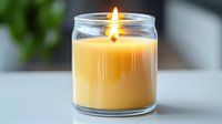 Burning yellow scented candle in clear glass jar with soft glowing flame against blurred background, creating cozy atmosphere for relaxation and aromatherapy.