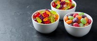 Colorful gummy bears and assorted candies in white bowls on dark slate surface for sweet treats and confectionery marketing materials.