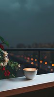 Cup of latte with heart-shaped foam art on balcony railing at dusk, alongside blooming flowers against blurred city lights background creating cozy atmosphere.