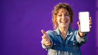 Happy young woman with curly hair showing smartphone screen and thumbs up gesture on purple background for marketing campaigns.