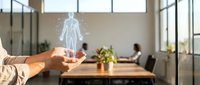 Businessman holding glowing human body hologram above wooden table in modern office with colleagues working in background, representing digital health technology and medical innovation.