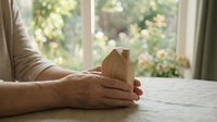 Mature hands holding wooden house model on table near window with garden view, representing real estate, property investment, home ownership, and housing market concepts.