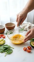 Hands whisking eggs in glass bowl surrounded by fresh vegetables including spinach, tomatoes and avocado for healthy breakfast preparation in modern kitchen.