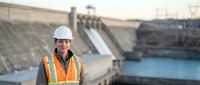Professional engineer in safety vest and hard hat standing at hydroelectric dam facility with concrete spillway and water reservoir in background.