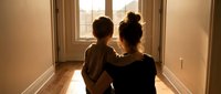 Mother and young son sitting together in sunlit hallway, embracing while looking out window. Warm family bonding moment capturing love and connection at home.