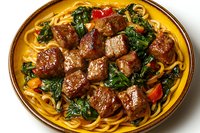 Tender beef chunks with sauteed kale and cherry tomatoes served over fresh linguine pasta on yellow ceramic plate, garnished with herbs and sauce.