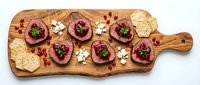 Gourmet beef carpaccio appetizer platter with pomegranate seeds, cheese, crackers and herbs on rustic wooden cutting board for fine dining presentation.