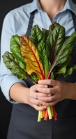 Person of indeterminate gender wearing light blue shirt and dark apron holding fresh rainbow chard bunch with vibrant green, yellow, and red stems for cooking, nutrition, and healthy eating concepts.