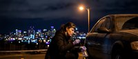 Young man sitting beside car at night using smartphone with city lights and street lamp creating atmospheric urban scene for lifestyle content.