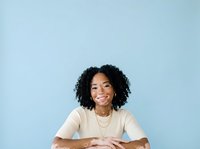 Young woman with dark curly hair and medium skin tone wearing beige t-shirt, smiling at camera with arms crossed against light blue background for portrait and lifestyle use.