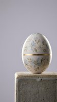 Elegant porcelain Easter egg with delicate floral pattern and gold trim displayed on concrete block against soft purple background for spring decor.
