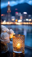 Romantic evening cityscape with glowing candle in crystal holder and white roses, blurred bokeh lights of urban skyline reflecting on water surface at dusk.