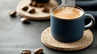 Steaming hot coffee in matte black ceramic cup on cork coaster with roasted coffee beans scattered around on dark textured surface, moody morning atmosphere.