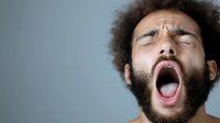 Young African American man with afro hair and beard yawning or expressing tiredness against gray background, showing natural emotion and fatigue.
